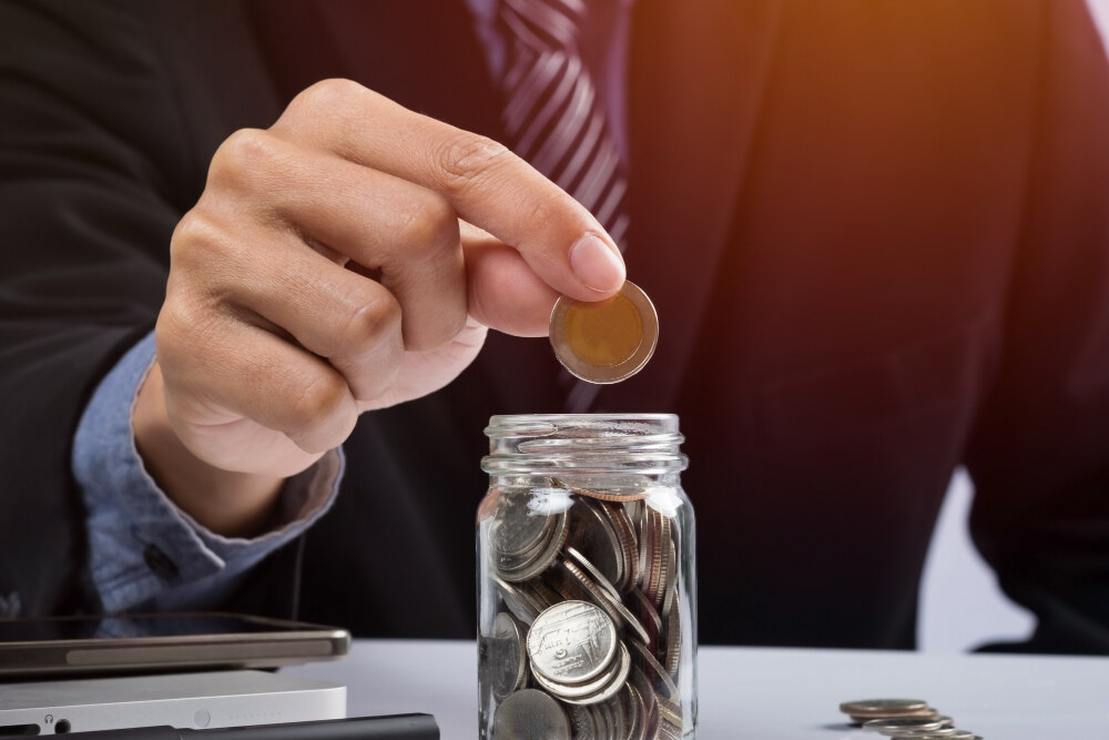 A businessman putting a coin into a money jar symbolizes investing in funds without a demat account.