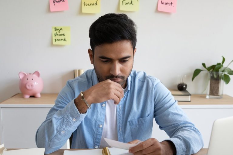 An Indian Man sitting at his desk with his laptop and papers, developing strategies for saving money on a 10,000 rupees income.