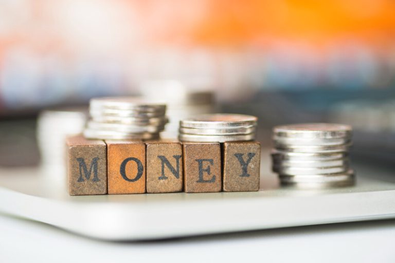 Wooden blocks spelling out the word "MONEY" with stacks of silver coins on a reflective surface, illustrating that financial security can be the best thing money can buy.